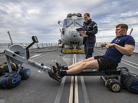man using rowing machine on deck being watched by colleague